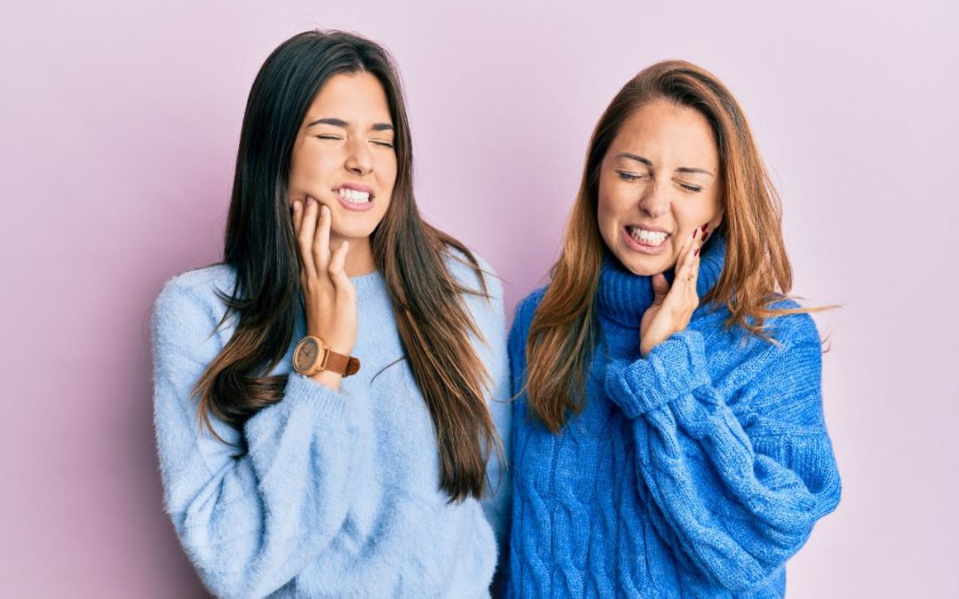 Two women in blue sweaters smiling and touching their faces against a pink background.